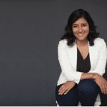 Author and Australian Writers’ Centre graduate Rashida Tayabali with white blazer sitting in front of blac background.