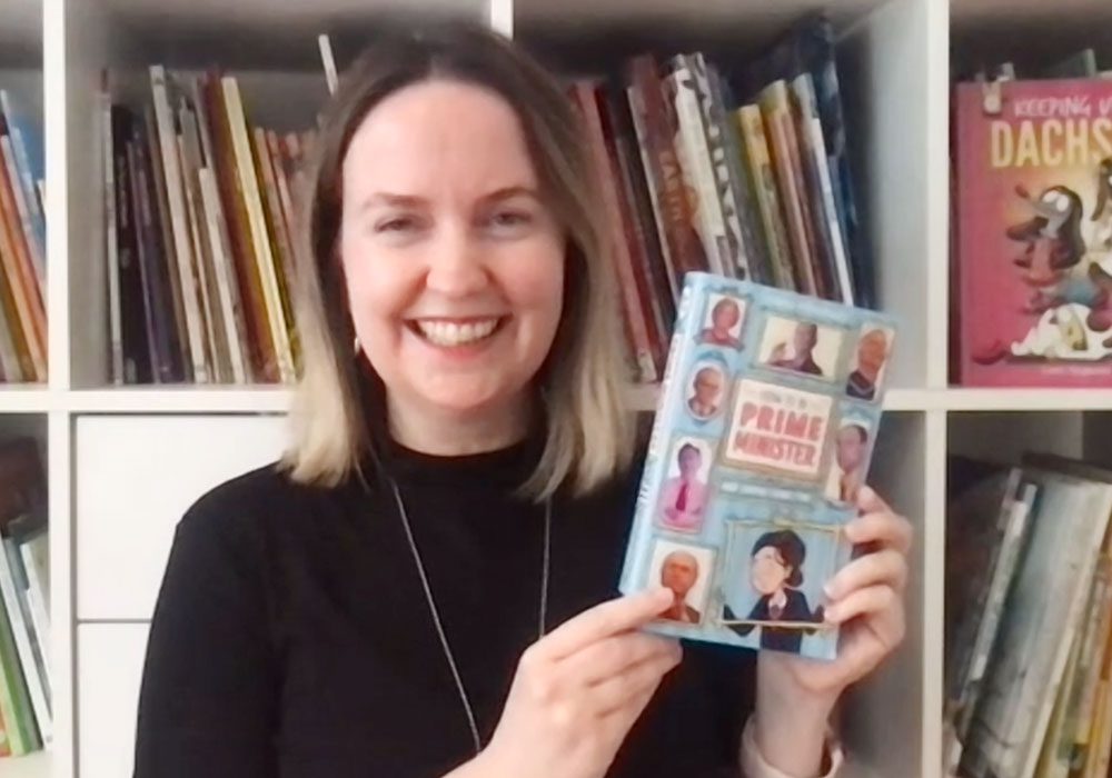 Author Carla Fitzgerald standing in front of a crowded bookcase, holding up a copy of her novel